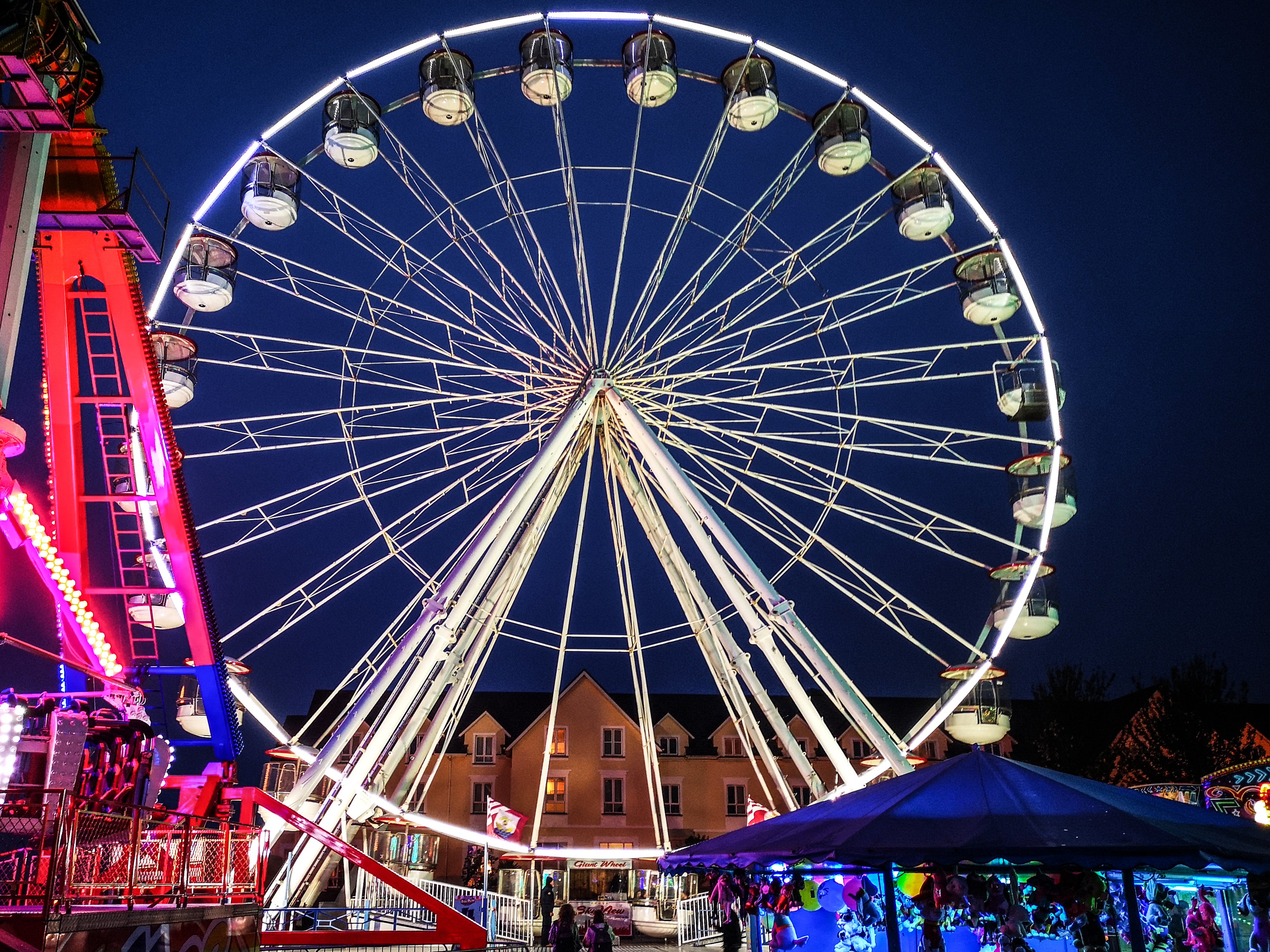 Grande ruota panoramica illuminata di notte in un luna park, con cabine vuote, giochi colorati in primo piano e palazzi residenziali sullo sfondo.