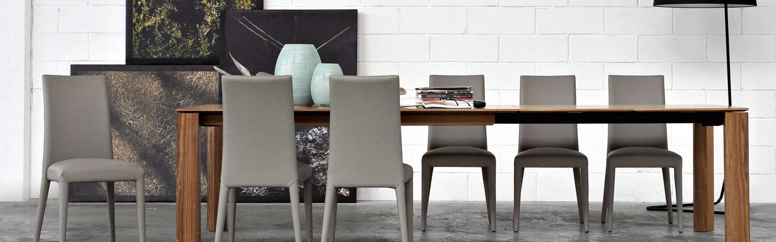 Tavolo da pranzo moderno in legno con sei sedie grigie, decorato con vasi ceramici azzurri e riviste, inserito in un ambiente industriale con pavimento in cemento e parete bianca a mattoni.