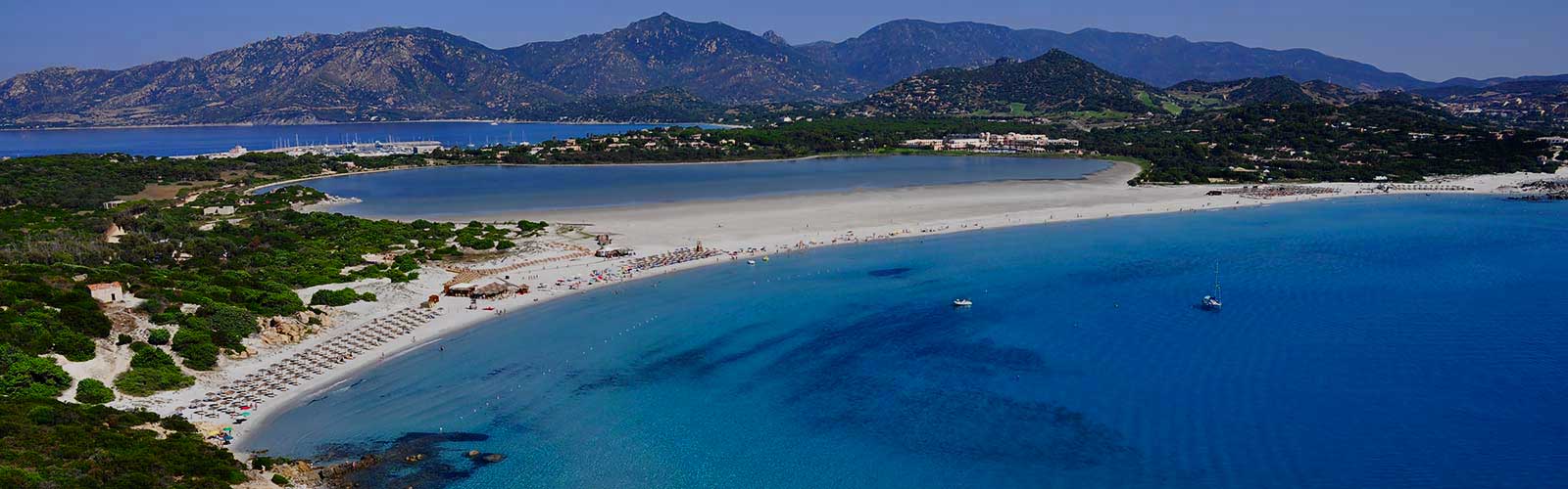 Veduta aerea della spiaggia di Porto Giunco, a Villasimius in Sardegna, con sabbia bianca, mare cristallino, ombrelloni disposti in fila e lo stagno retrostante, circondato da vegetazione e montagne sullo sfondo.