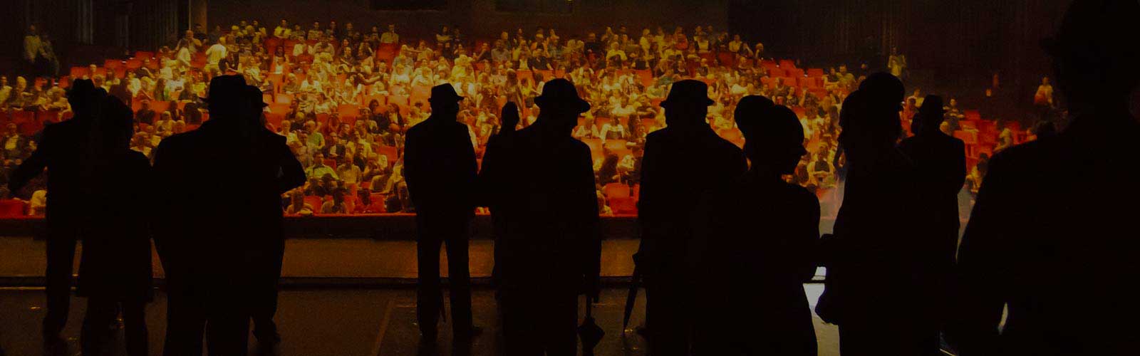 Persone in controluce viste dal palco di un teatro affollato, rivolte verso una platea illuminata piena di spettatori seduti.
